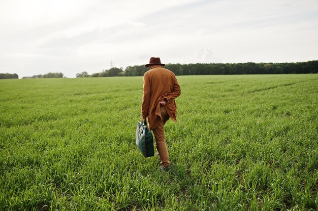 Stylish man in glasses brown jacket and hat with bag posed on green field