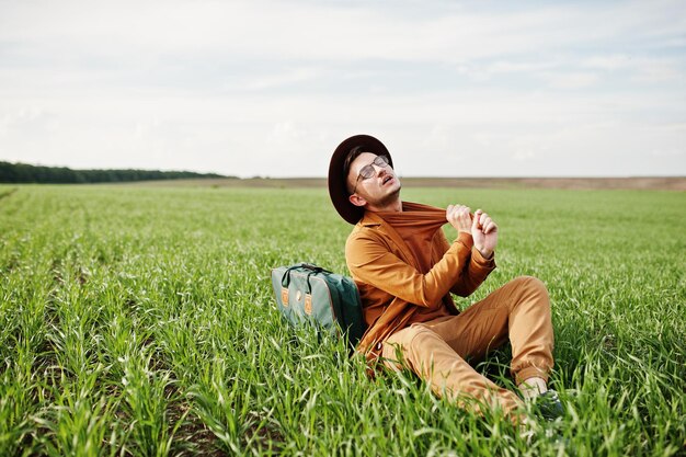 Stylish man in glasses brown jacket and hat with bag posed on green field