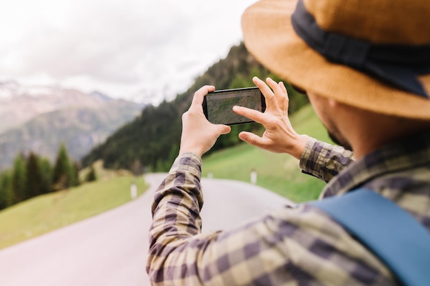 Stylish man has lost his way and looking for new route on map in the phone walking down the road. Outdoor portrait from back of traveler in checkered shirt going to mountains in vacation.