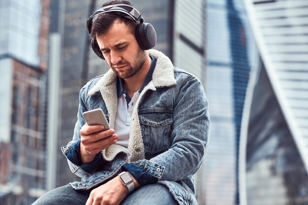 曇りの朝にモスクワ市の高層ビルの前に座って音楽を聴き、スマートフォンを使用してデニムジャケットを着ているスタイリッシュな男。
