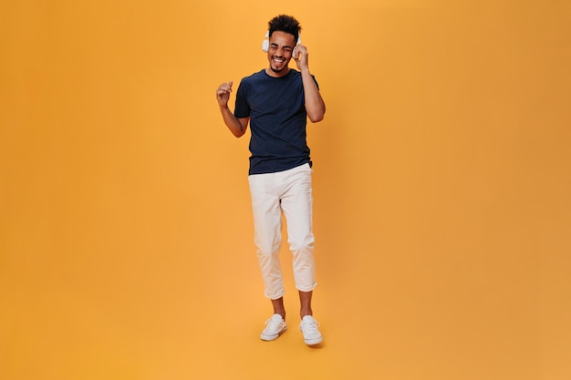 Стильный мужчина в белых штанах слушает музыку в массивных наушниках Портрет парня в темно-синей футболке, танцующего на оранжевом фоне