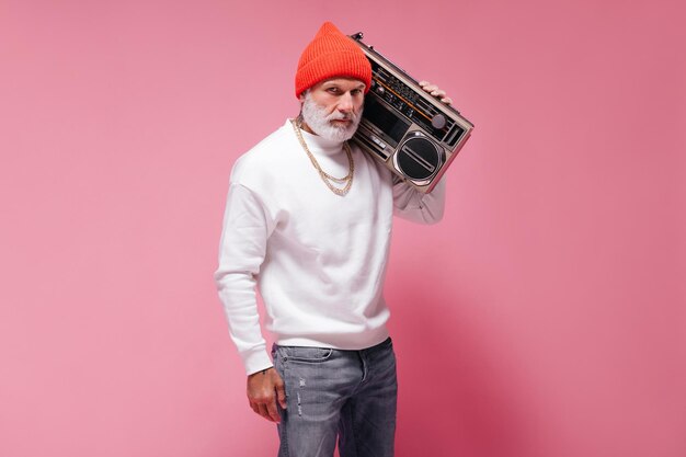 Stylish man in white sweater posing with record player Serious bearded guy in orange hat with gold chains looking into camera