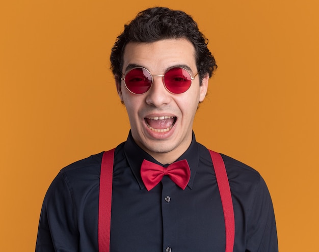 Free photo stylish man with bow tie wearing glasses and suspenders happy and excited looking at front standing over orange wall