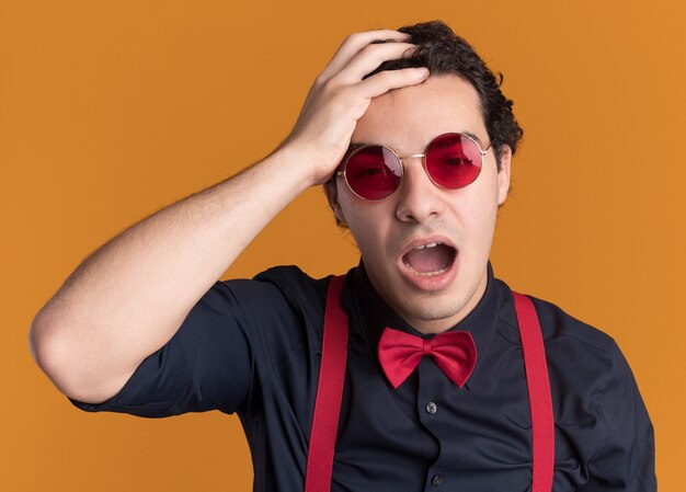 Stylish man with bow tie wearing glasses and suspenders looking confused with hand on his head for mistake standing over orange wall