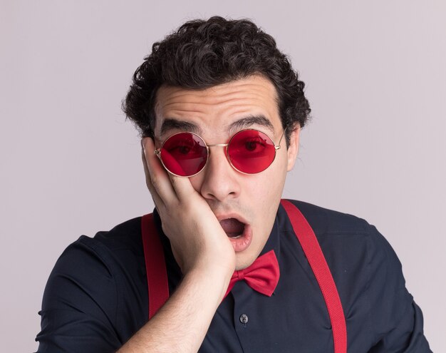 Stylish man with bow tie wearing glasses and suspenders looking at front amazed and surprised standing over white wall