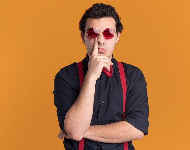 Stylish man with bow tie wearing glasses and suspenders looking at front being confused and displeased standing over orange wall