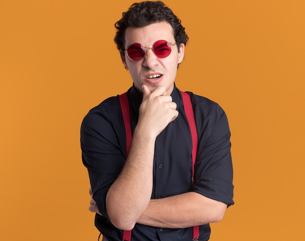 stylish man with bow tie wearing glasses and suspenders looking at front confused with hand on his chin standing over orange wall