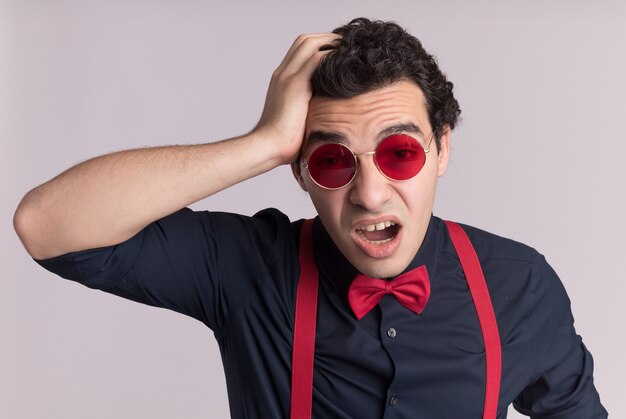 Stylish man with bow tie wearing glasses and suspenders looking at front confused with hand on his head for mistake standing over white wall