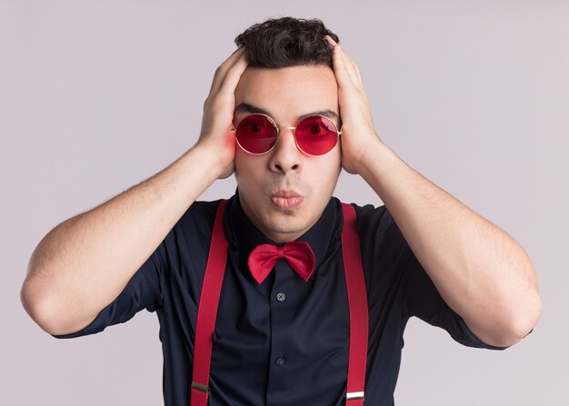 Stylish man with bow tie wearing glasses and suspenders looking at front confused and worried with hands on his head standing over white wall