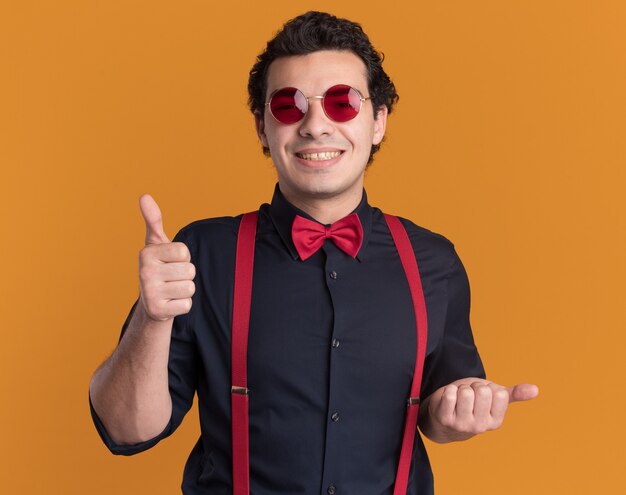 안경과 멜빵을 착용하는 나비 넥타이와 세련된 남자가 오렌지 벽 위에 서서 행복하고 긍정적 인 보여주는 엄지 손가락을 웃고 있습니다.