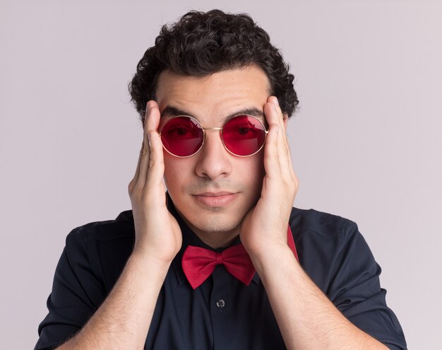 Stylish man with bow tie wearing glasses and suspenders looking at front tired and bored touching his temples standing over white wall