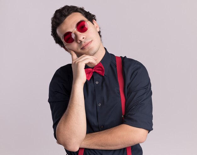 Stylish man with bow tie wearing glasses and suspenders looking at front with pensive expression thinking standing over white wall