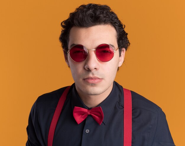 Stylish man with bow tie wearing glasses and suspenders looking at front with skeptic expression standing over orange wall