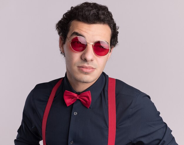 Stylish man with bow tie wearing glasses and suspenders looking at front with skeptic expression standing over white wall