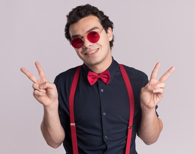 Stylish man with bow tie wearing glasses and suspenders looking at front with smile on face showing v-sign standing over white wall