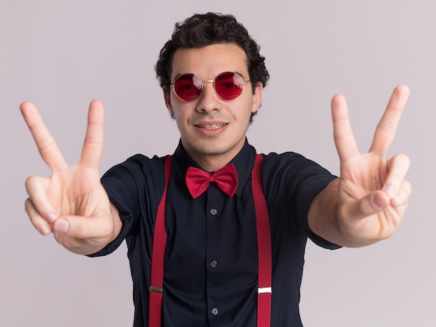 Free photo stylish man with bow tie wearing glasses and suspenders looking at front with smile on face showing v-sign standing over white wall