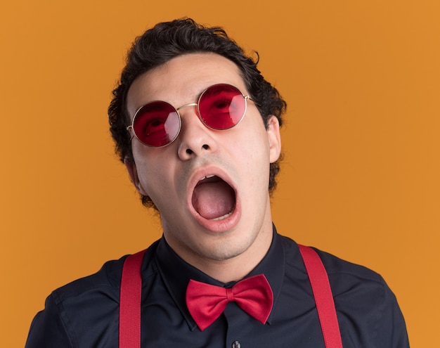 Stylish man with bow tie wearing glasses and suspenders looking up with wide opened mouth standing over orange wall