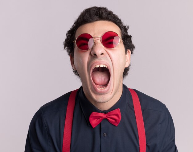 Stylish man with bow tie wearing glasses and suspenders shouting with aggresive expression going wild standing over white wall