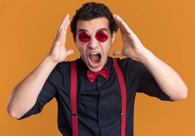 Free photo stylish man with bow tie wearing glasses and suspenders shouting with arms raised going wild standing over orange wall