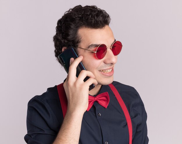 안경과 멜빵을 착용하는 나비 넥타이와 세련된 남자가 흰 벽 위에 서있는 휴대 전화로 이야기 웃고