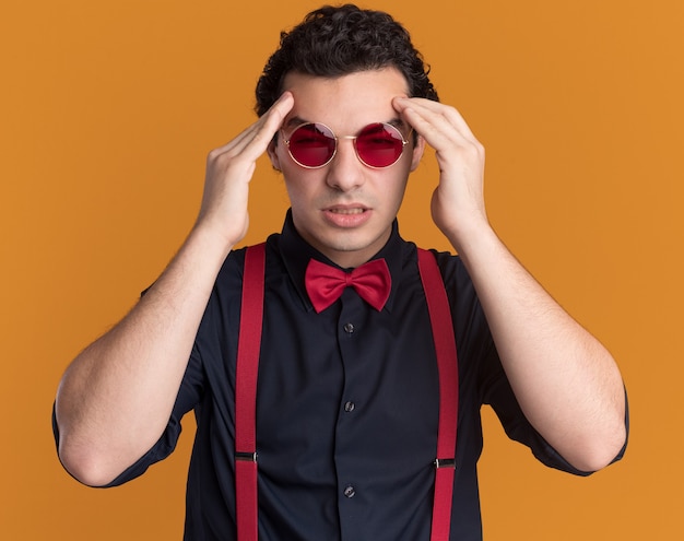 Uomo alla moda con il farfallino con gli occhiali e le bretelle che tocca la sua testa alla ricerca di mal di testa che soffre di mal di testa in piedi sopra la parete arancione
