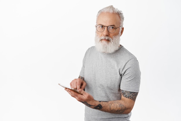 Stylish mature bearded man with tattoos, reading in glasses, using digital tablet and looking at front serious, standing in gray t-shirt against white wall