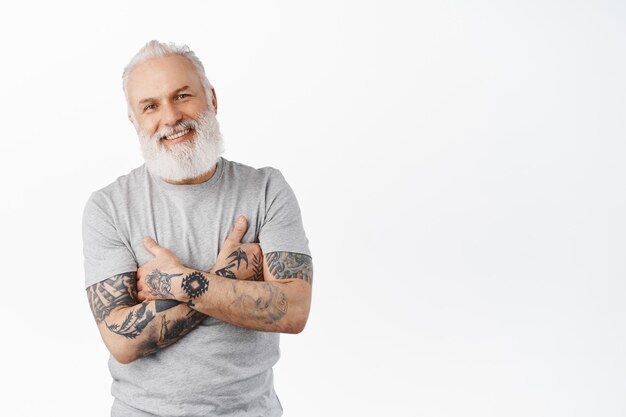 Stylish mature man with tattoos and long beard cross arms on chest, smiling and looking candid at camera. Authentic carefree senior man standing against white background
