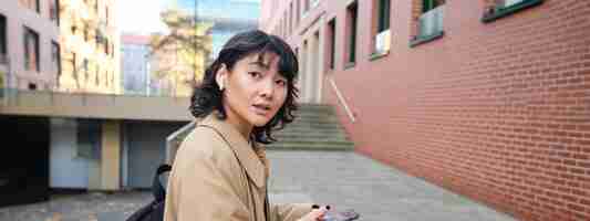 無料写真 ワイヤレスヘッドフォンを着たスタイリッシュな現代アジアの女子学生が笑顔でバックパックを持って街を歩き回る
