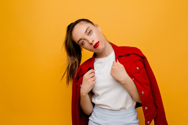 Stylish modern female model dressed red jacket, white t-shirt and blue skirt with red lipstick poses on isolated wall. Fashion, style, look, model, place for text