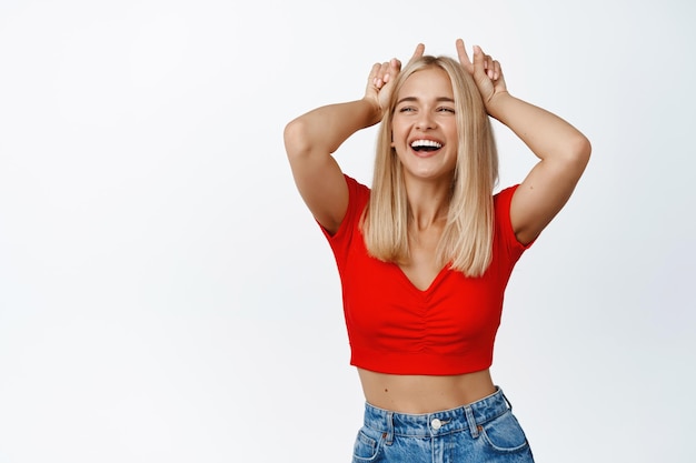 Stylish modern girl dancing with bull devil horns gesture fingers on head smiling and laughing carefree white background