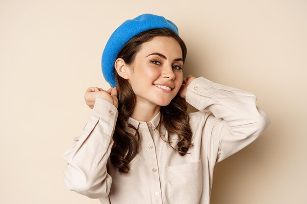 Stylish modern girl put on trendy hat on head and smiling going out posing against beige background