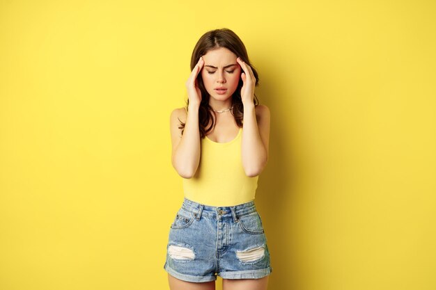 Stylish modern woman rubbing temples, having headache, migraine, frowning and grimacing from pain in head, standing against yellow background