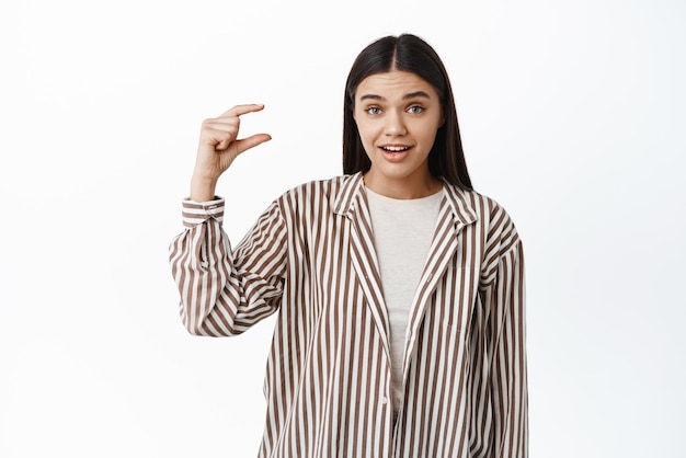 Stylish modern woman showing something small making little size gesture on copy space staring at camera surprised standing over white background