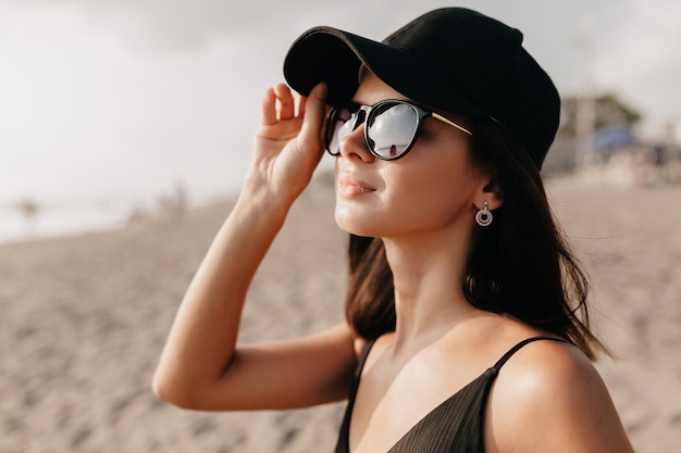 Elegante donna moderna in abito di tendenza guardando sull'oceano con un sorriso felice che indossa cappello e occhiali e gode di calde giornate estive giovane modello femminile caucasico in riva al mare