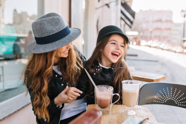 Стильная мама и довольно улыбающаяся дочь наслаждаются выходными вместе в ресторане под открытым небом, пьют кофе и молочный коктейль.