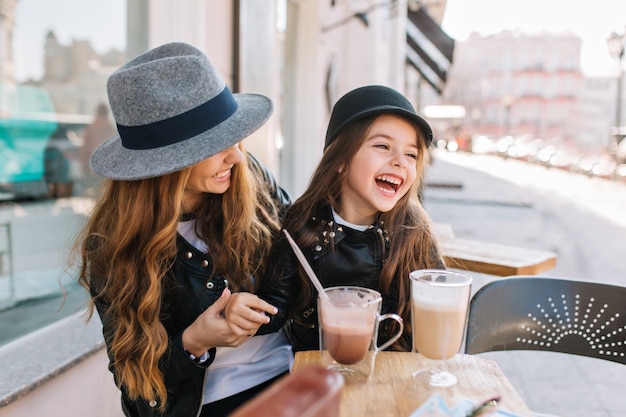 세련된 엄마와 예쁜 웃는 딸이 커피와 밀크 쉐이크를 마시는 야외 레스토랑에서 주말을 함께 즐기고 있습니다.