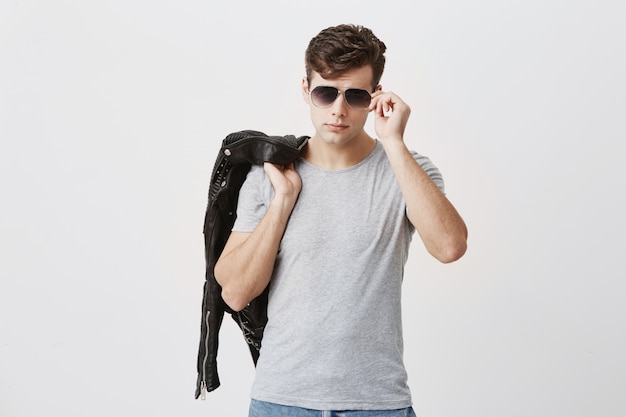 Stylish, muscular, confident male posing . Attractive handsome caucasian guy with trendy haircut with black leather jacket thrown over shoulder, holding sunglasses in hand