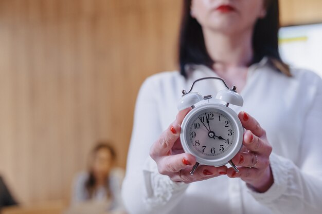 Stylish office worker woman in glasses with a classic alarm clock in the hands on background of working colleagues
