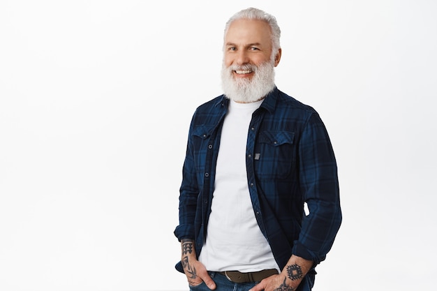 Stylish old man with beard and tattoos, standing relaxed with hands in pockets, looking aside at logo banner with pleased smile, standing over white wall