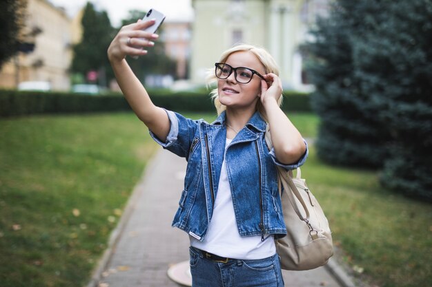 Stylish pretty fashion blonde girl woman in jeans suite makes selfie on her phone in the city in the morning