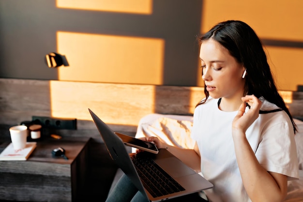 Stylish pretty lady with long brown hair in headphones is sitting in the bed in sunlight in morning and working on laptop and holding smartphone in the hands