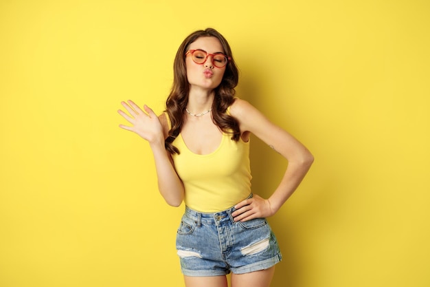 Stylish pretty woman posing in sunglasses and summer clothes, looking happy and carefree, concept of vacation and sunny days, standing over yellow background