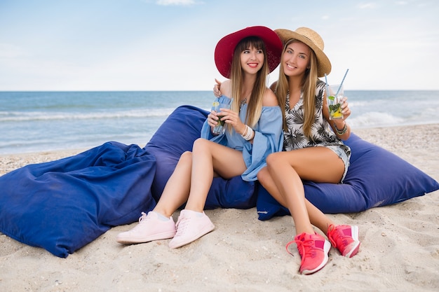 Stylish pretty women on summer vacation on tropical beach, friends together, fashion trend accessories, smiling, skinny legs, sitting on sand, having fun long legs in sneakers