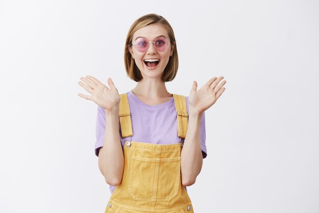 Stylish pretty young woman in sunglasses and summer clothes looking carefree, clasp hands and looking excited, rejoicing with great news