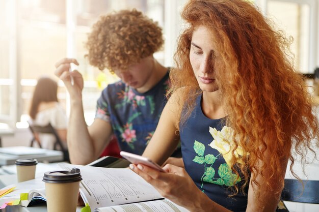 Stylish red-haired female receiving a message from her friend while studying at coffee shop