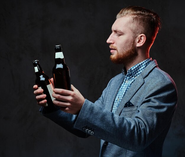 Stylish redhead bearded male dressed in a suit holds a bottle with craft beer over grey background.