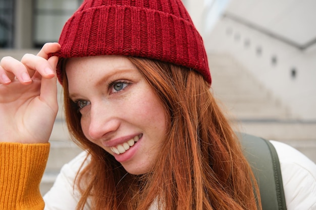 Бесплатное фото Стильная рыжая девушка в теплой красной шляпе улыбается расслабленно, сидя с рюкзаком на лестнице возле здания w