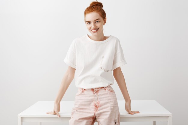 Stylish redhead girl lean on table and standing over white wall