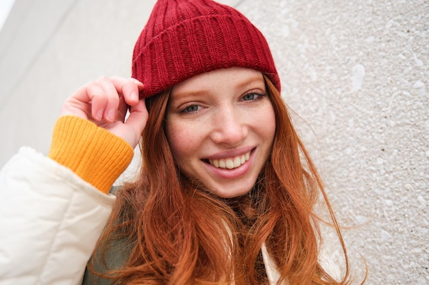 Free photo stylish redhead girl in red hat smiles and looks happy poses outdoors on street looks relaxed and li