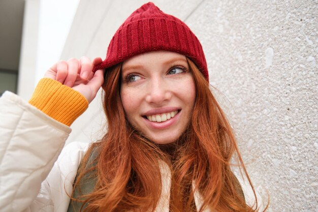 Стильная рыжая девушка в красной шляпе улыбается и выглядит счастливой, позирует на улице, выглядит расслабленной и жизнерадостной.